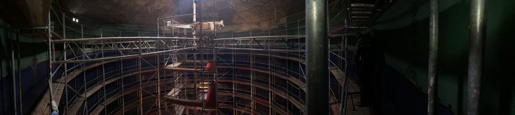 Weitwinkelaufnahme aus dem Inneren der Biogasanlage Güterglück, mit einem Metallgerüst und einer nach oben führenden Leiter. Die gedämpfte Beleuchtung hebt die geschwungenen Wände und die Tragkonstruktion hervor und lässt auf laufende Aktivitäten wie Wartungs- oder Bauarbeiten schließen.